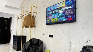 A modern hotel room with a large TV displaying various game shows, a black sofa, a round wooden table with a plant, and a gold-colored hanging chair. - ESE luxury spa suites (Rome)