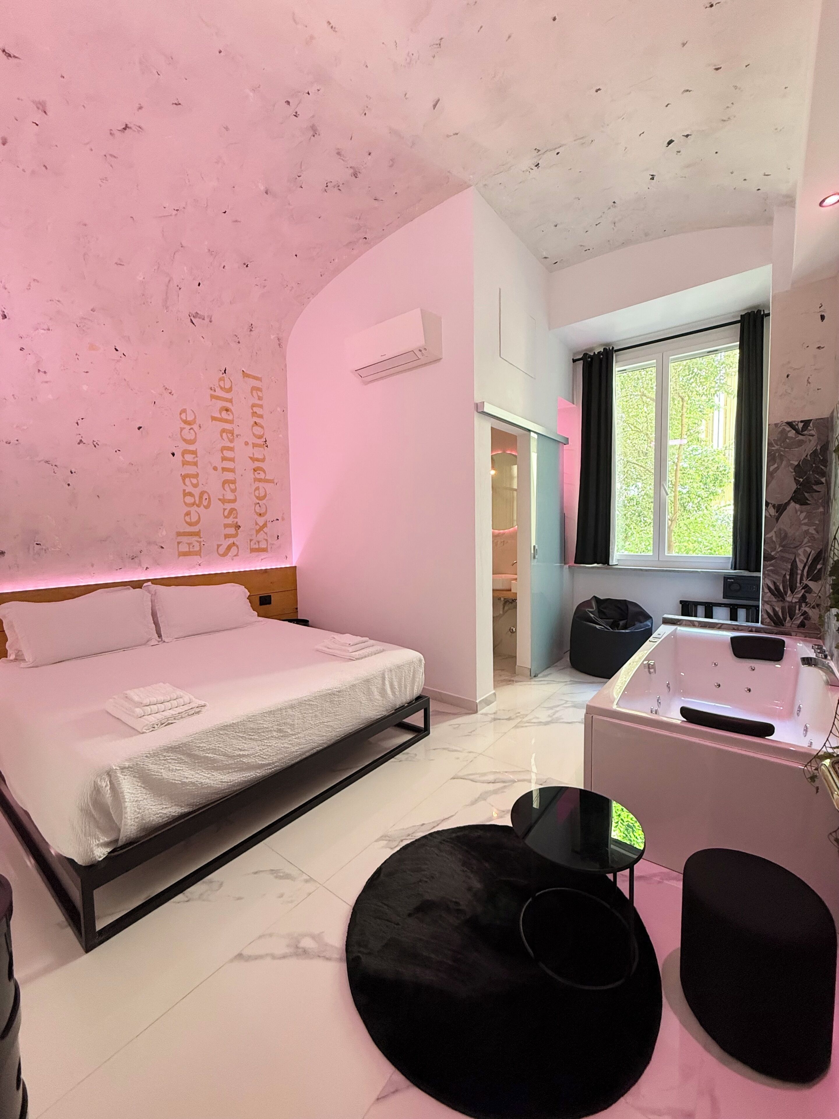 Een slaapkamer met een bed, een zwart poufje, een tafeltje, een raam met gordijnen en een badkamer die door een geopende deur zichtbaar is.