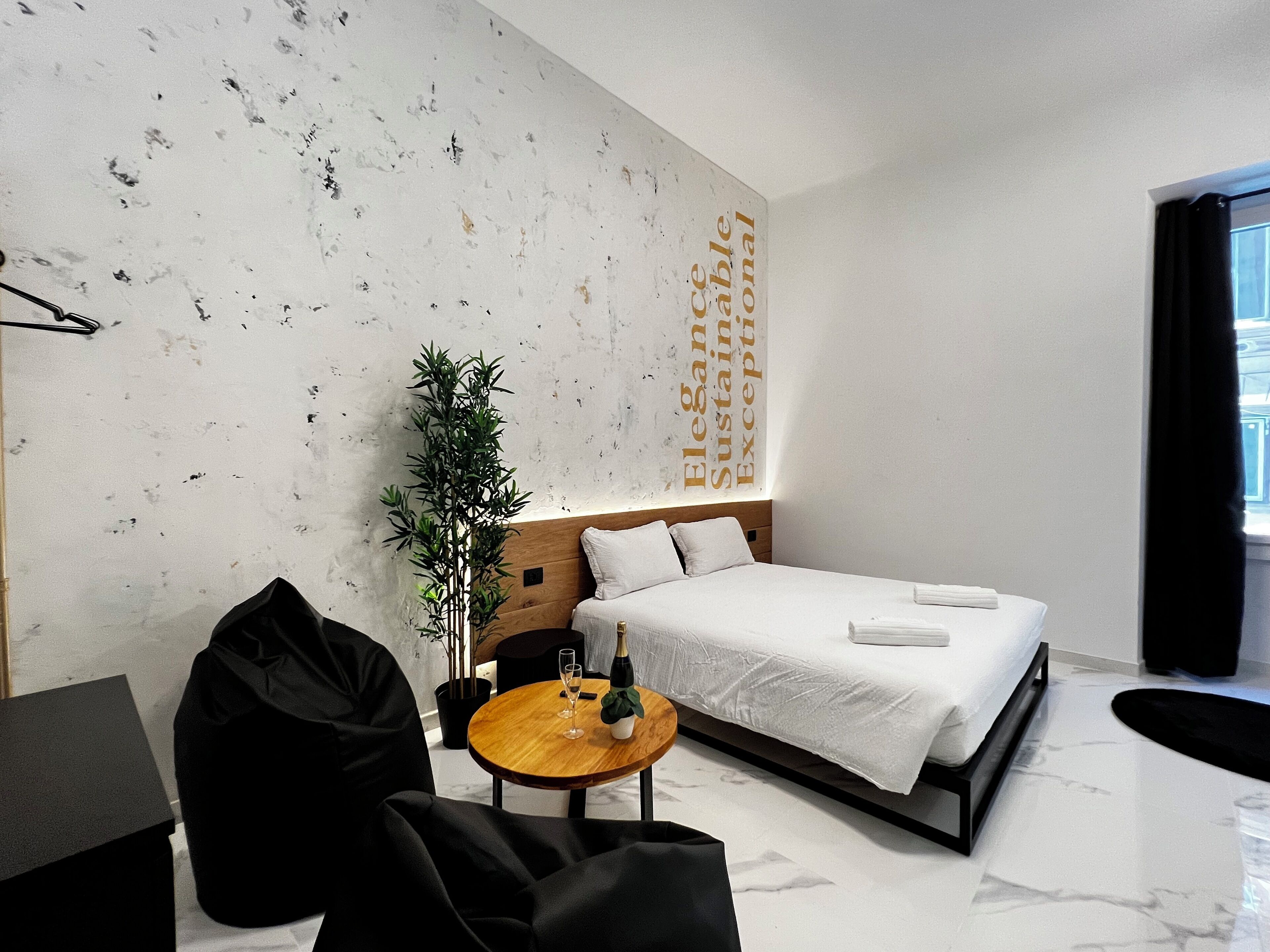 A modern hotel room with a large bed, a small round table, a potted plant, and a wall with a motivational quote.