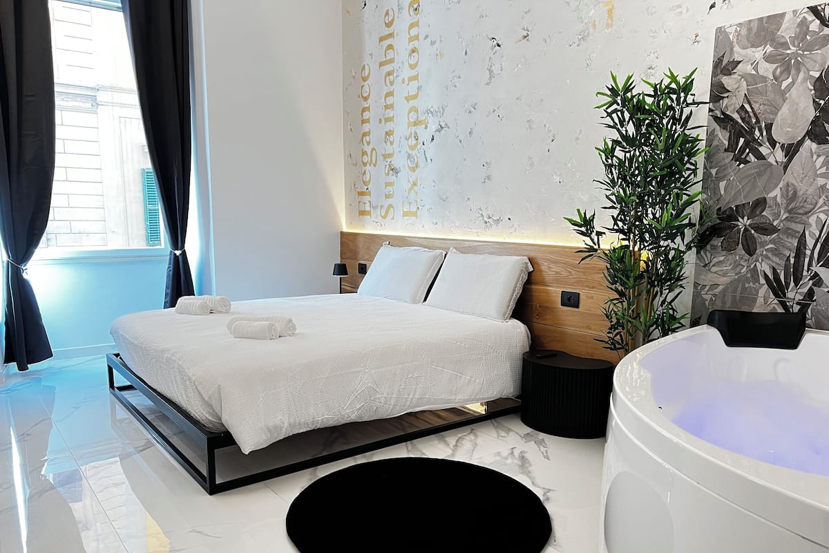 A modern bedroom with a bed, a black round rug, a large bathtub, and a potted plant.
