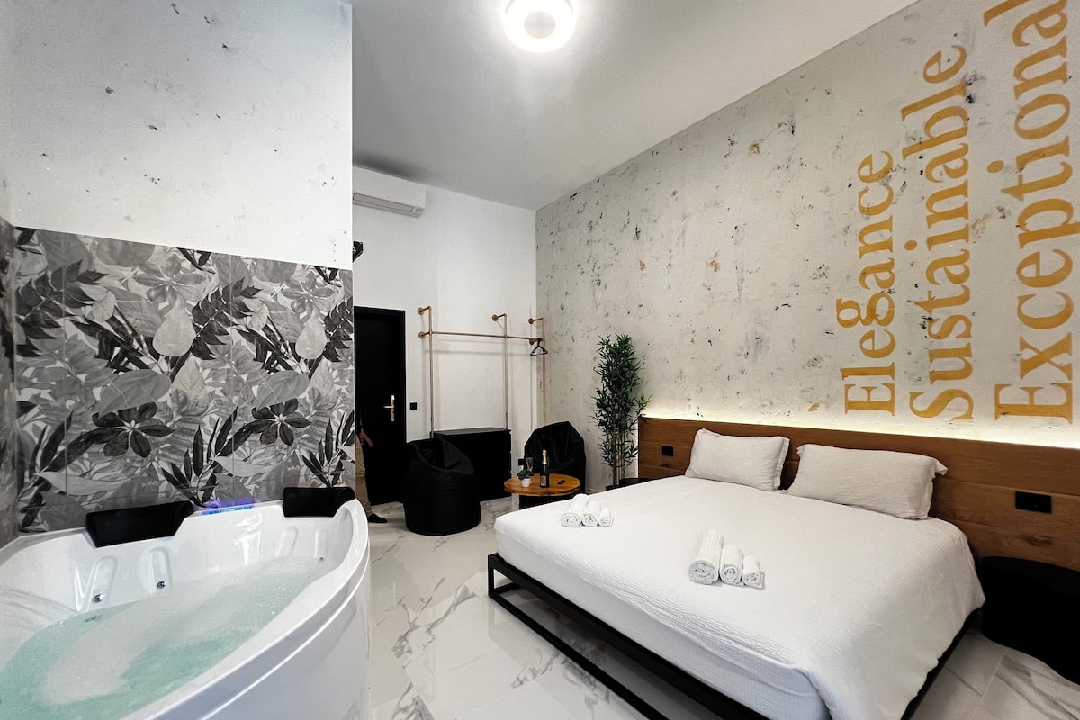 A modern hotel room with a large bed, a bathtub, and a wall with the words "Elegance Sustainable Exceptional".