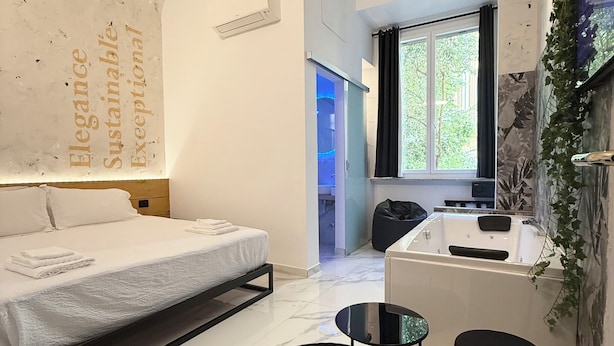 A modern hotel room with a bed, a small table, a black ottoman, a bathtub, and a window with a view of greenery. - ESE luxury spa suites (Rome)