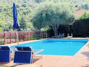 Una piscina rectangular con agua azul cristalina, rodeada por una terraza con baldosas. Hay dos sillones azules y una sombrilla. Un árbol grande proporciona sombra a la derecha de la piscina.