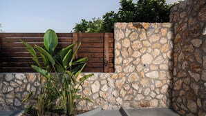 A stone wall with a wooden fence, a concrete planter with green plants, and a stone wall with a metal fixture. - Kathys Apartment 2 in Archaggelos (Αρχαγγελος)