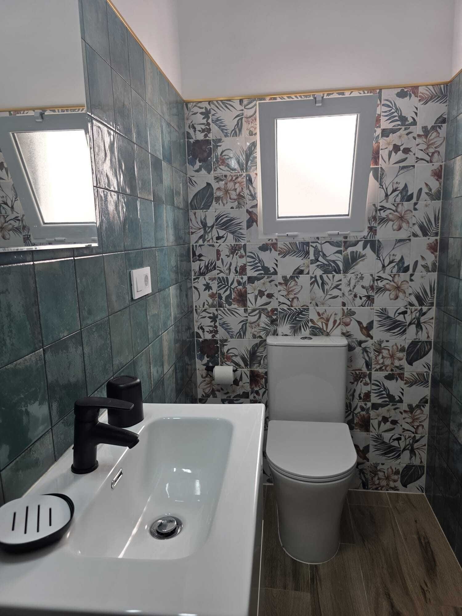A bathroom with a white sink, black faucet, and a toilet. There is a window above the toilet and a mirror above the sink. The walls are tiled with a floral pattern.
