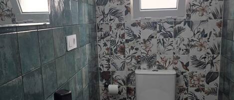 A bathroom with a white sink, black faucet, and a toilet. There is a window above the toilet and a mirror above the sink. The walls are tiled with a floral pattern.