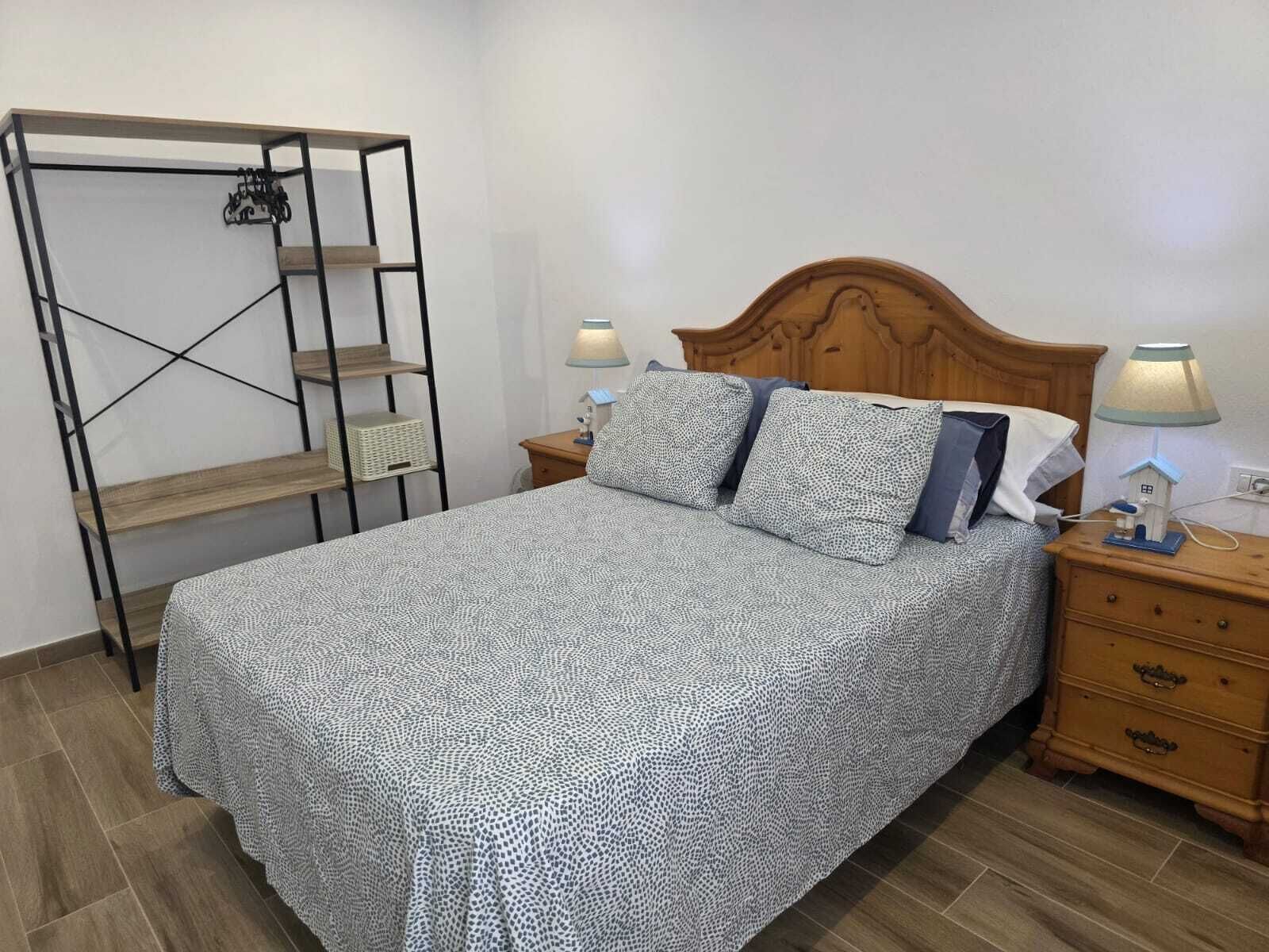 A bed with a patterned bedspread, two bedside lamps, a wooden nightstand, a wooden headboard, and a metal shelving unit with a basket.