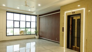 A spacious lobby with a large window, a potted plant, a ceiling fan, and an elevator. - The Crown Leela (Pratapgarh)