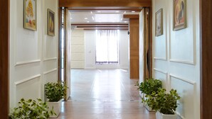 A hallway with wooden flooring, white walls, and ceiling lights. There are potted plants on either side and framed pictures on the walls. - The Crown Leela (Pratapgarh)