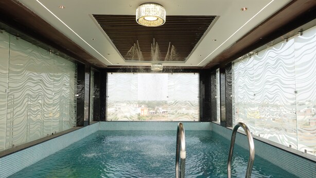 An indoor swimming pool with a view of a cityscape through a large window. - The Crown Leela (Pratapgarh)