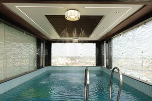 An indoor swimming pool with a view of a cityscape through a large window. - The Crown Leela (Pratapgarh)