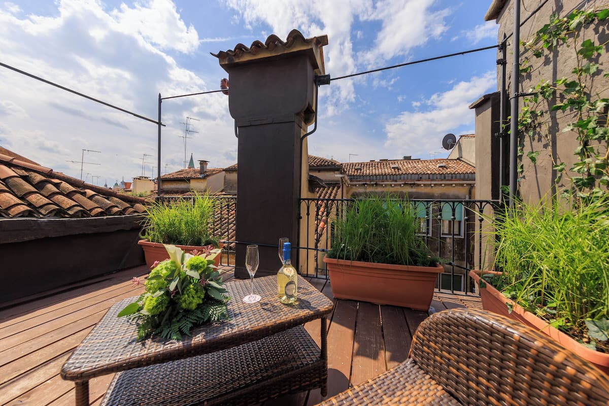 A rooftop terrace with a wicker table, potted plants, and a bottle of wine.