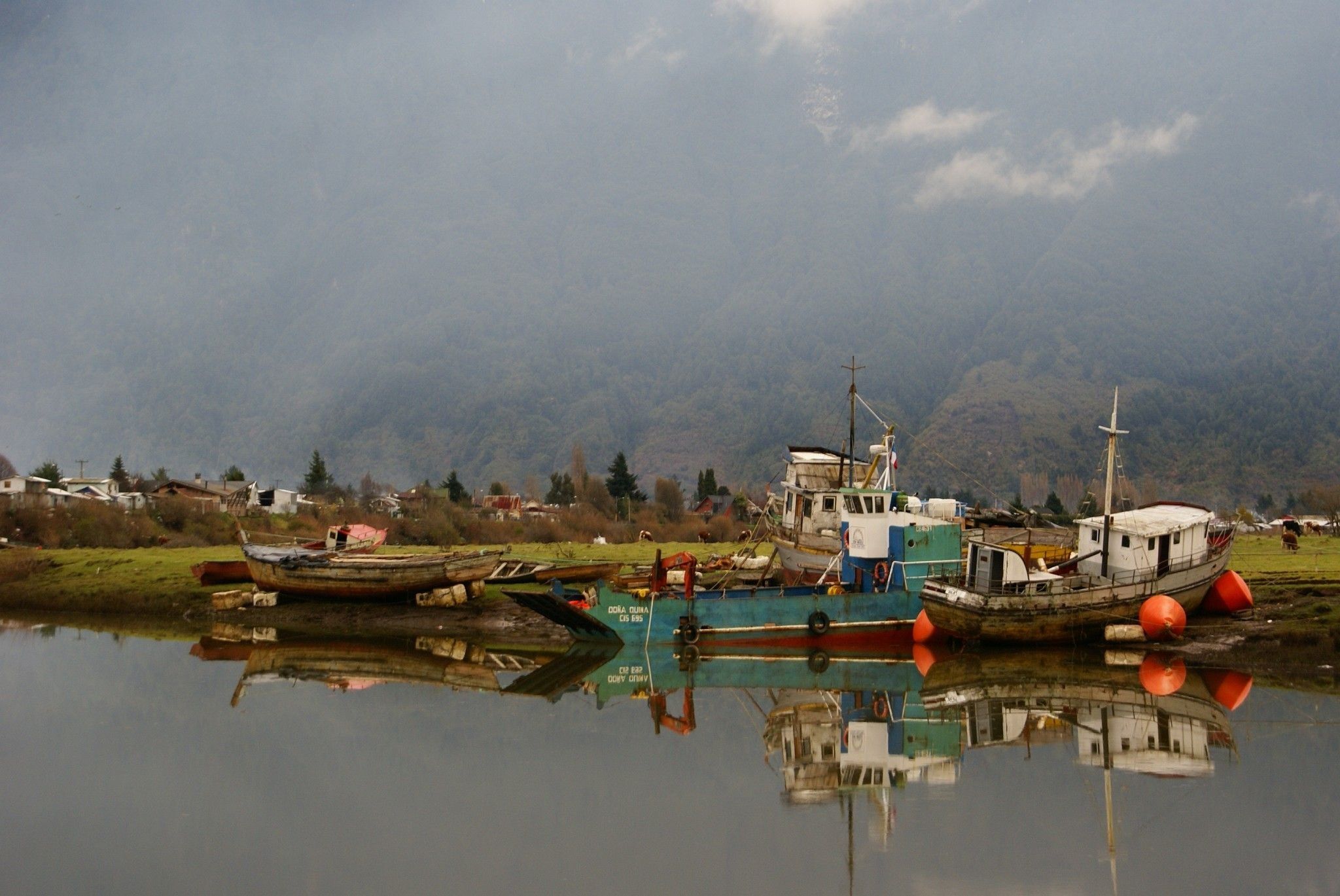 Une rivière paisible où sont amarrés plusieurs bateaux, le tout entouré d’un paysage montagneux.