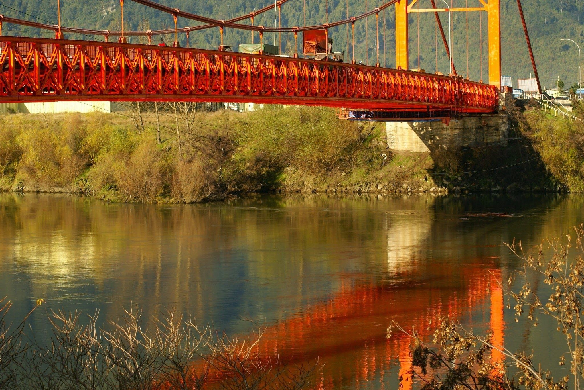Un puente rojo sobre un río tranquilo con una ladera boscosa al fondo.