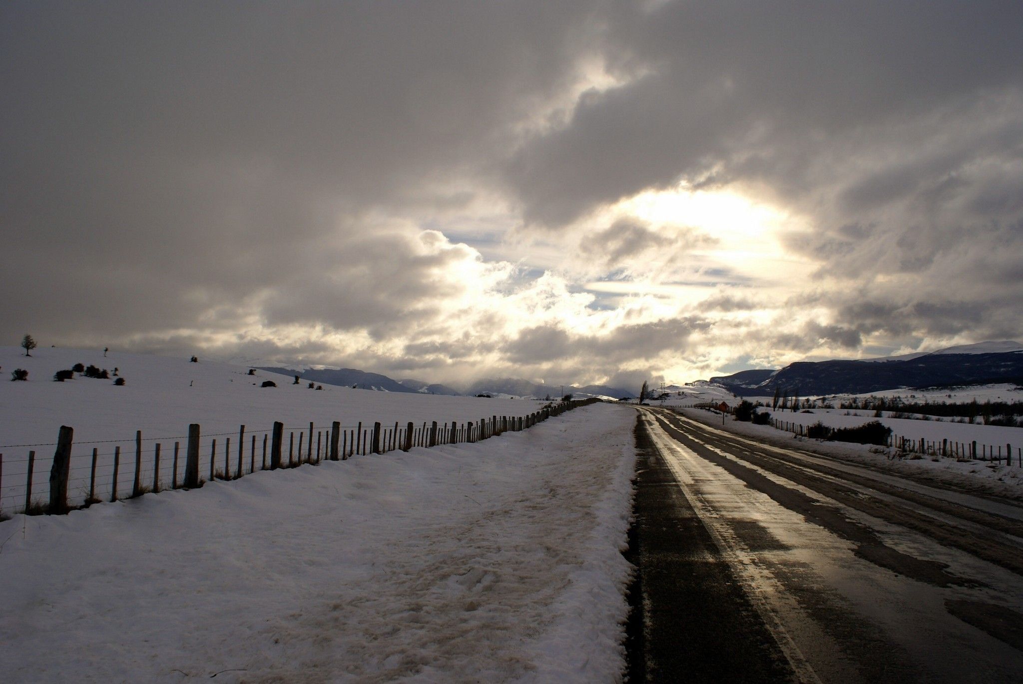 A snow-covered road with a fence on one side and a mountain range in the distance.