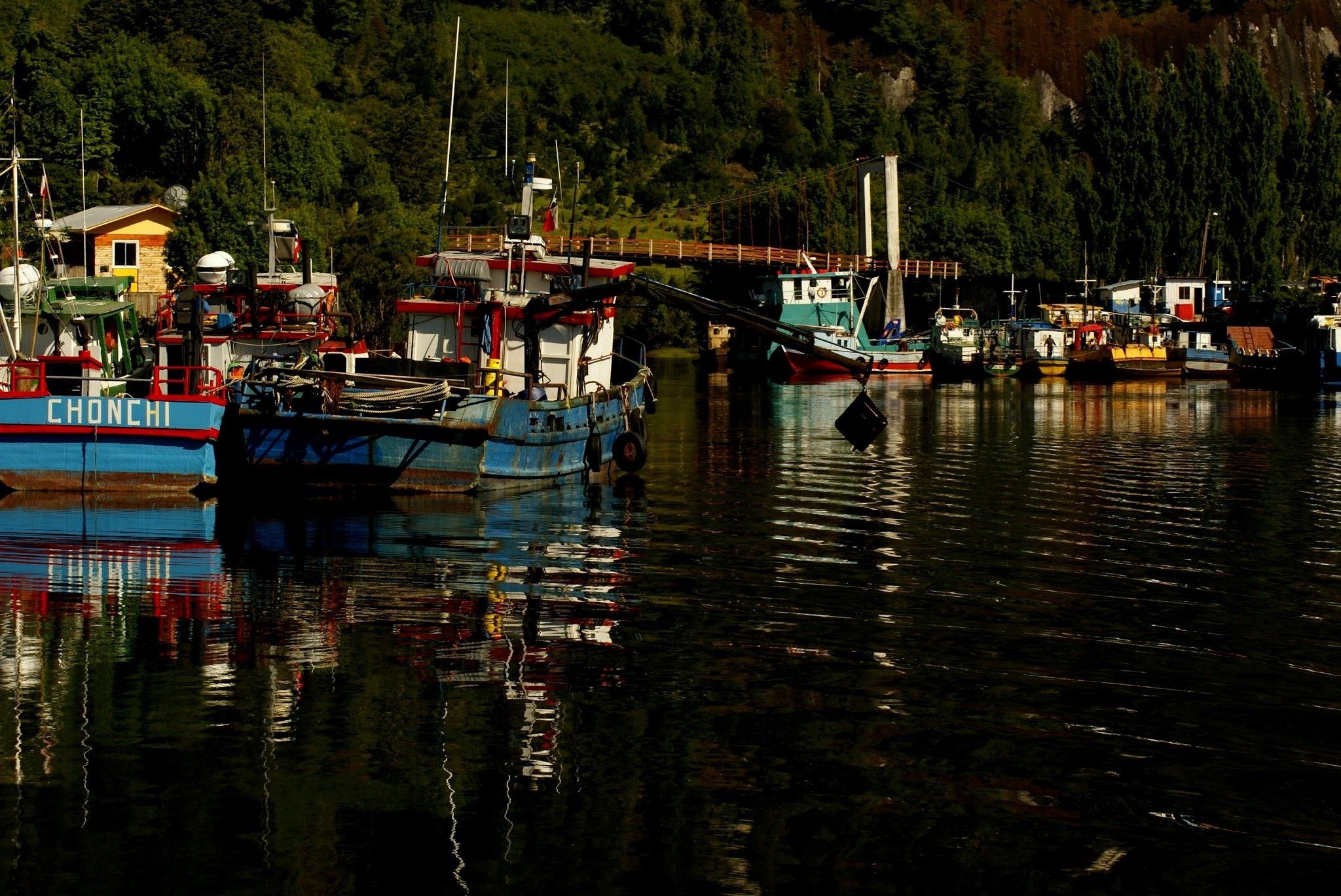 Ein Hafen mit vertäuten Booten, einer Brücke und einer bewaldeten Hügelkette im Hintergrund.