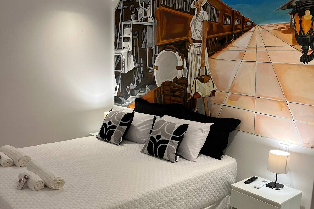Una habitación de hotel moderna con una cama grande, una mesita de noche, una lámpara y un mural en la pared.