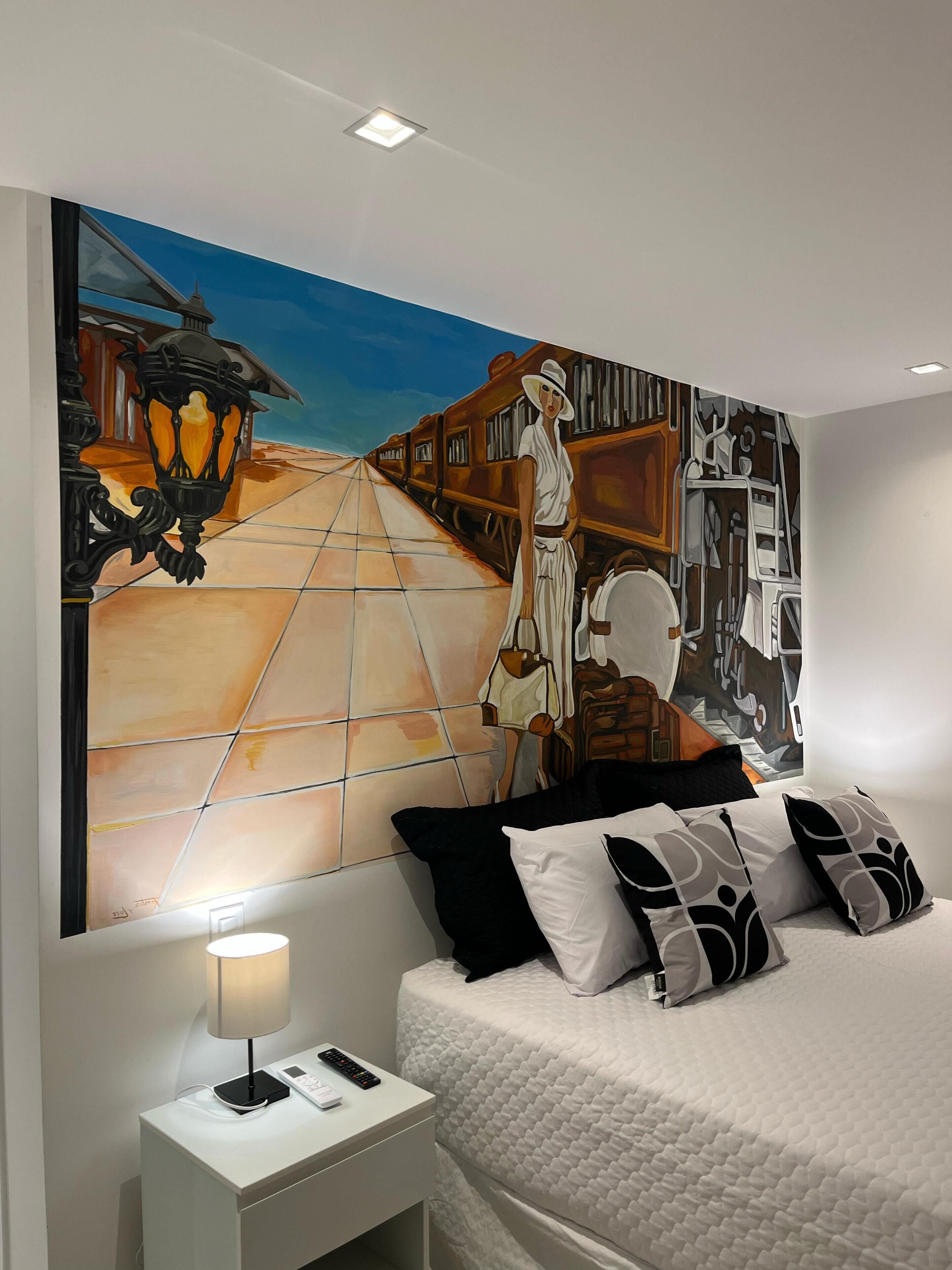 A bedroom with a large abstract painting on the wall, a bed with white bedding and patterned pillows, a bedside table with a lamp, and a nightstand with a remote control.