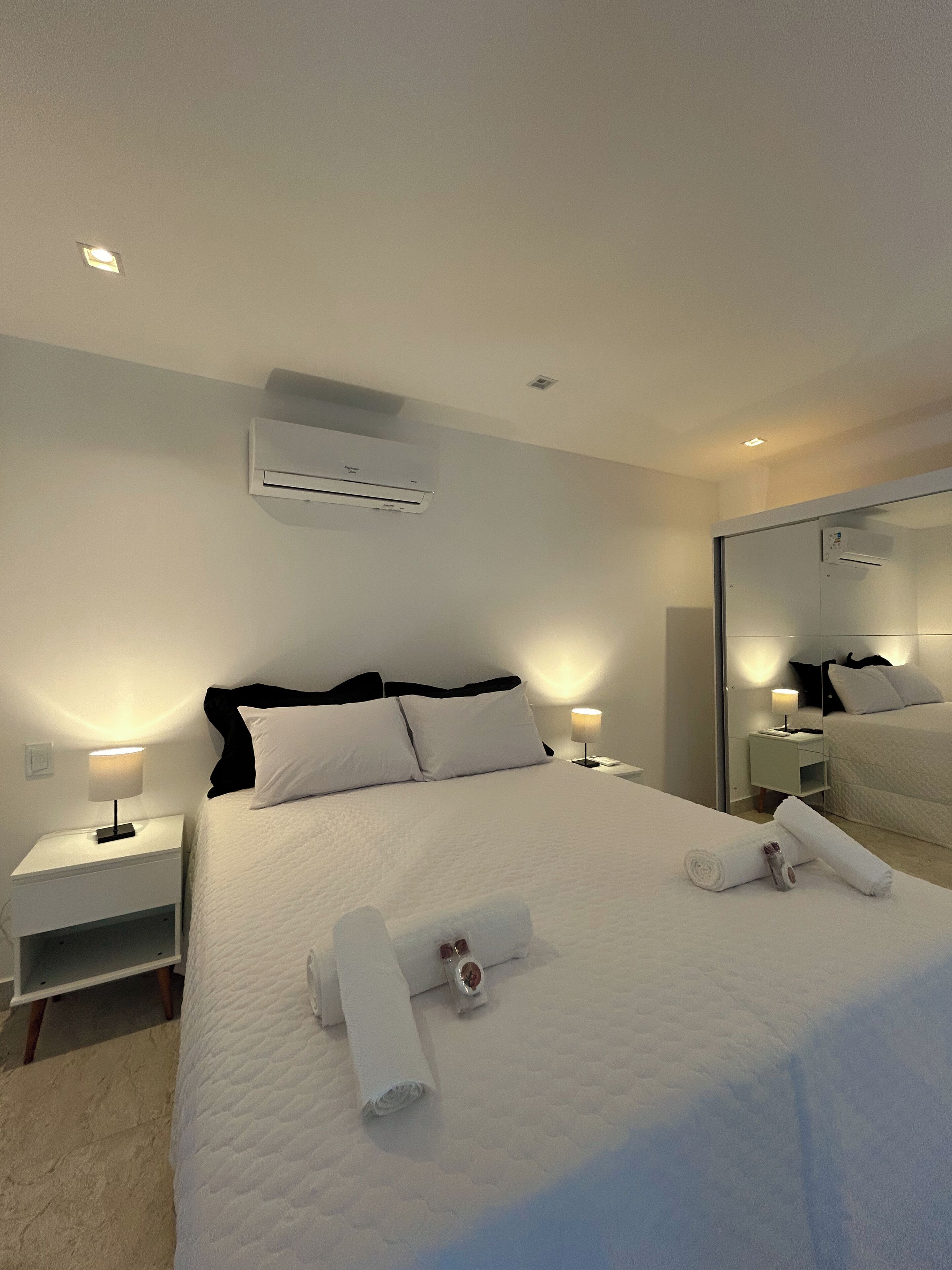 Habitación de hotel con una cama bien hecha, dos mesitas de noche con lámparas, aire acondicionado instalado en la pared y un espejo grande.