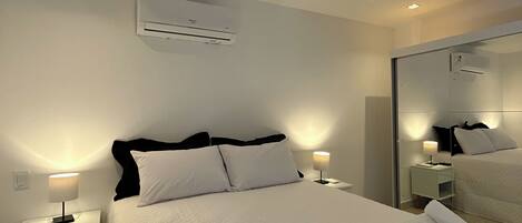 Habitación de hotel con una cama bien hecha, dos mesitas de noche con lámparas, aire acondicionado instalado en la pared y un espejo grande.