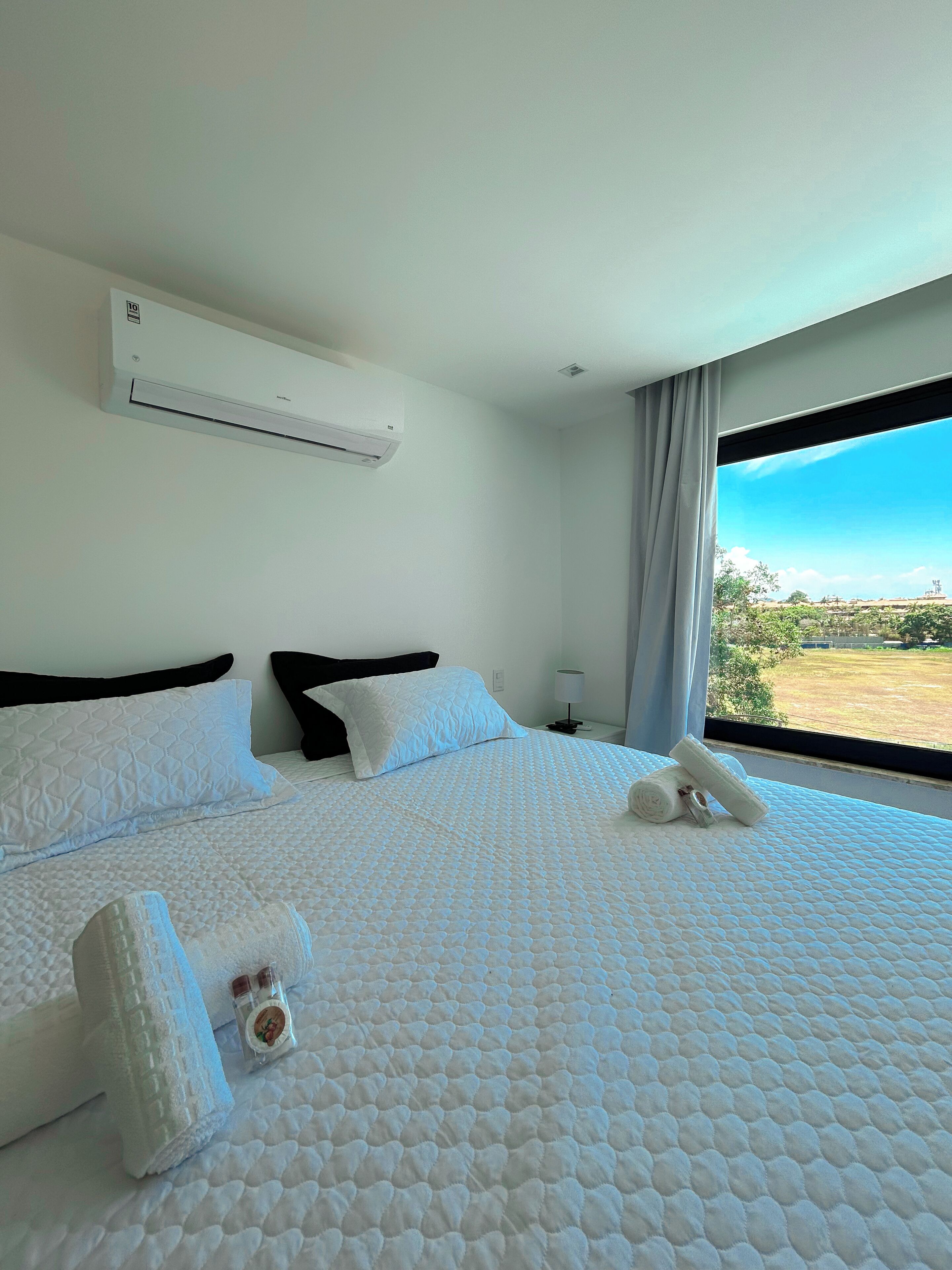 A hotel room with a large bed, white bedding, a window with a view of the countryside, and an air conditioning unit on the wall.