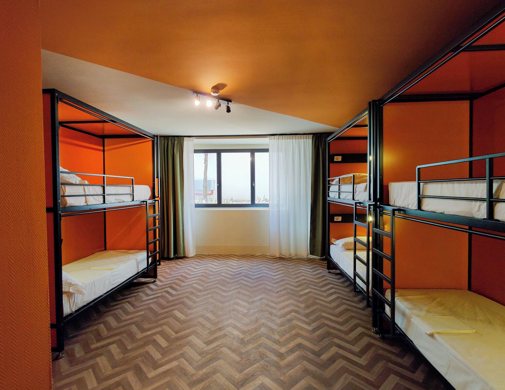 A dormitory room with bunk beds, a window with curtains, and a herringbone-patterned floor.