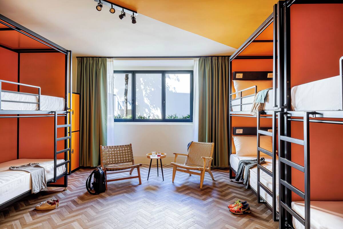 Une chambre de dortoir avec des lits superposés, deux fauteuils en osier, une petite table et une fenêtre avec des rideaux.