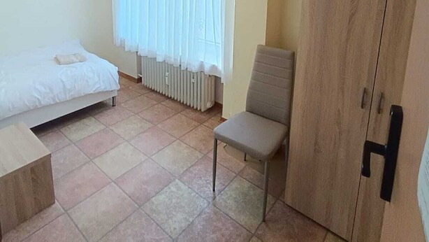 Ein kleiner, gut beleuchteter Raum mit einem Bett, einem Sessel und einem Kleiderschrank.