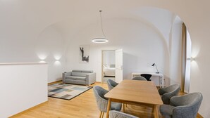 A modern, open-plan living and dining area with a wooden table, grey upholstered chairs, a grey sofa, and a patterned rug. - Hotel Benedict (Koszeg)