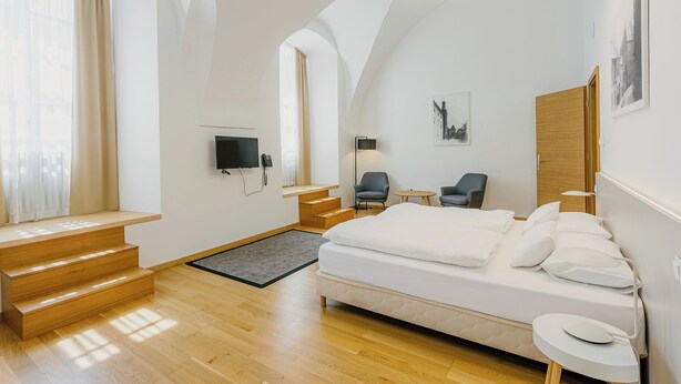 A modern bedroom with a large bed, a desk, a chair, a TV, and a staircase. - Hotel Benedict (Koszeg)