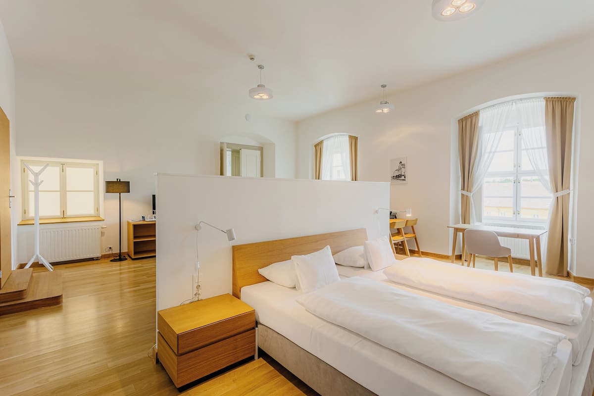 Habitación de hotel moderna con una cama grande, un escritorio y una silla. Cuenta con una ventana con cortinas, un radiador y una lámpara.