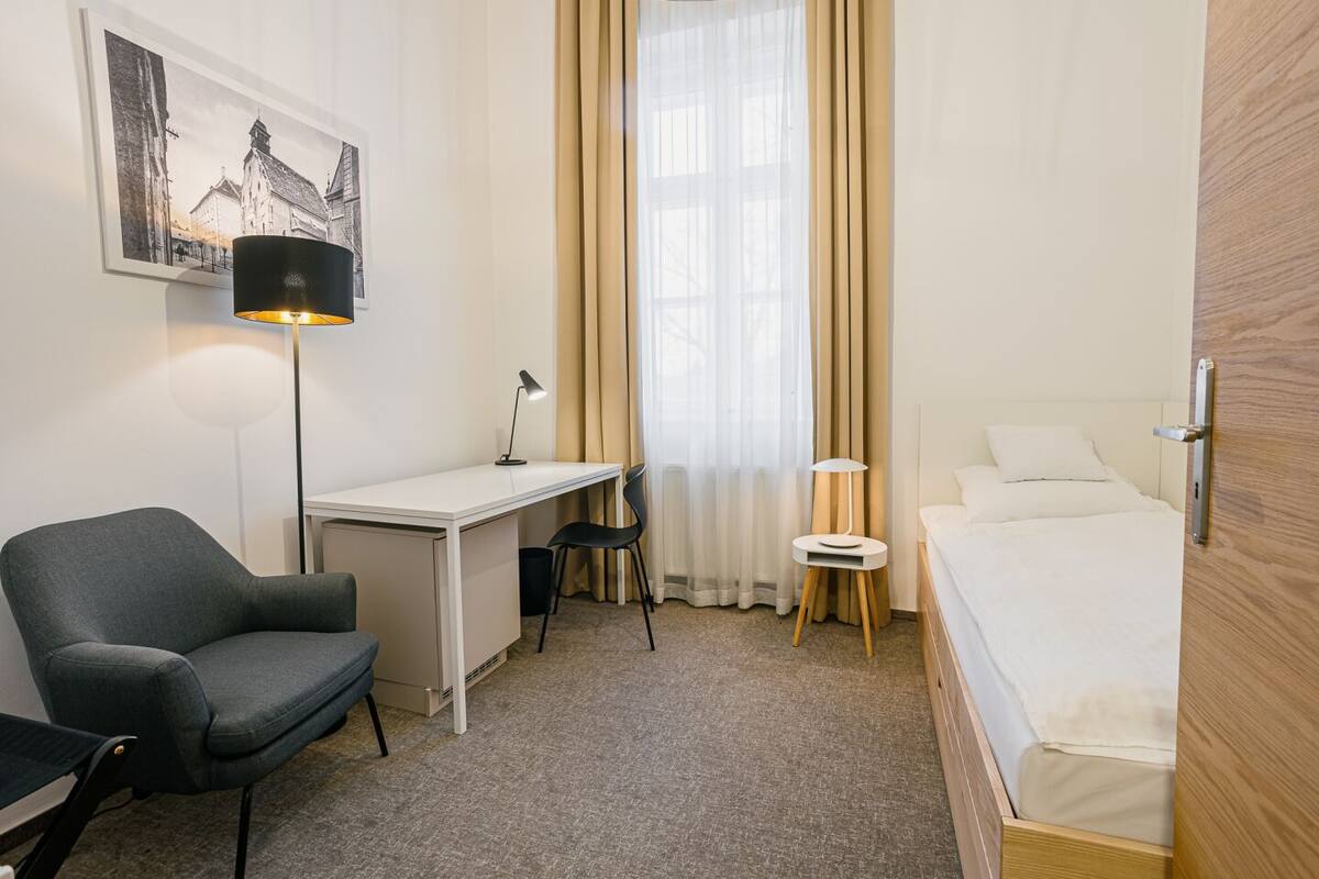 Habitación de hotel con cama, escritorio, silla, lámpara, un cuadro en la pared y ventana con cortinas.
