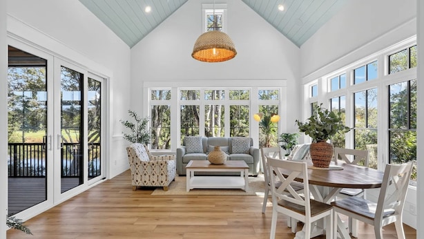 A spacious room with a high ceiling, large windows, a wooden floor, a sofa, a dining table with chairs, and a hanging light fixture. - Luxury Palmetto Dunes Home w/ Private Pool (Hilton Head Island)