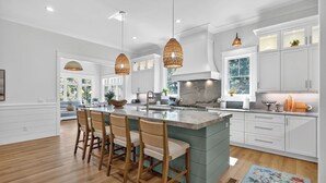 A modern kitchen with white cabinetry, a large island with bar stools, pendant lighting, and a view of greenery outside. - Luxury Palmetto Dunes Home w/ Private Pool (Hilton Head Island)