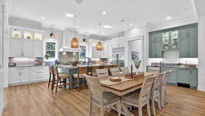 A modern kitchen with white cabinets, a central island with seating, and a dining area with a wooden table and chairs. - Luxury Palmetto Dunes Home w/ Private Pool (Hilton Head Island)