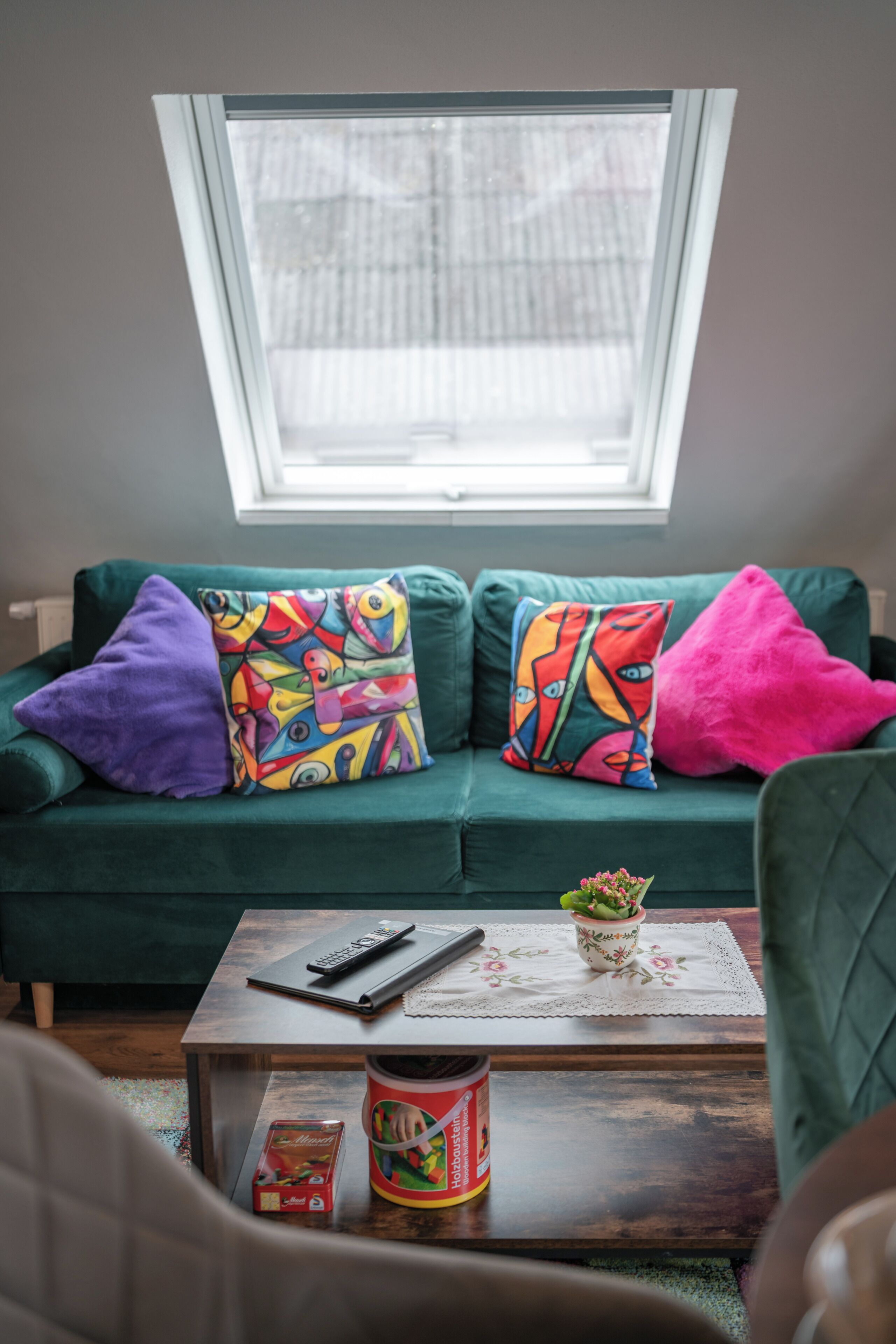 A green sofa with colorful pillows, a remote control, a book, and a can of coffee on a wooden table.