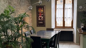 A dining area with a table and chairs, a potted plant, a framed picture on the wall, and a window with curtains. - Spacious apartment in cévenol village house (Barre-des-Cévennes)