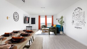 A modern living room with a dining area, featuring a wooden table set for four, a TV mounted on the wall, and a large window with orange curtains. - *NEW* The Boho Bungalow | 20%  Discount | BOOK NOW (New Braunfels)