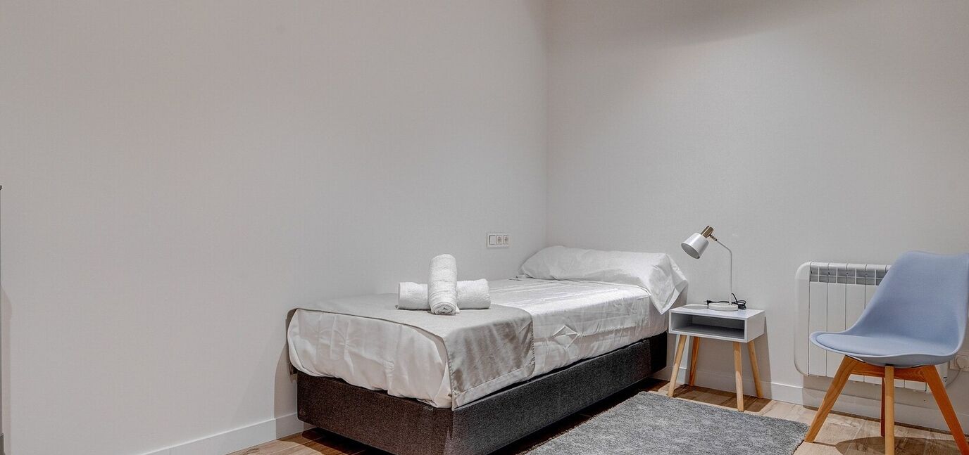 A bedroom with a bed, a nightstand, a chair, a lamp, and a radiator.
