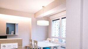 Une pièce moderne et bien éclairée, avec une table et des chaises blanches, une étagère intégrée ornée d’un vase, et une fenêtre donnant sur des immeubles.