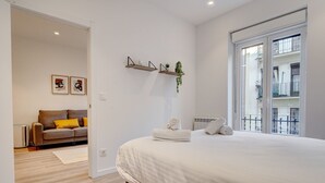 A bedroom with a large bed, a window with a view of a balcony, a small sofa with cushions, and a wall-mounted shelf with decorative items.