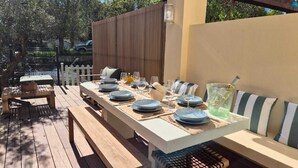 Un espace de restauration en plein air avec une longue table en bois pouvant accueillir six personnes, des bancs et un coin salon confortable agrémenté de coussins à rayures.