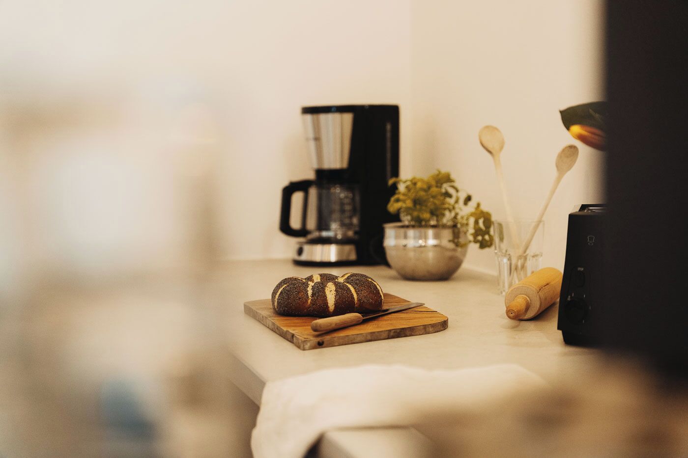 Eine Küchenarbeitsplatte mit Brot, einer Kaffeemaschine, einer Schüssel mit Besteck und einer Pflanze.
