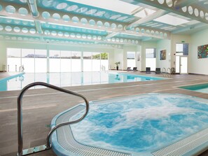A large indoor swimming pool with a hot tub, clear windows, and a modern ceiling design.