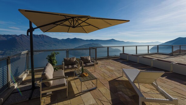 A terrace with outdoor seating, a table with a bottle, and a view of mountains and water.