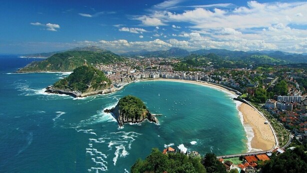An aerial view of a coastal city with a beach, a bridge, and a mountainous backdrop.