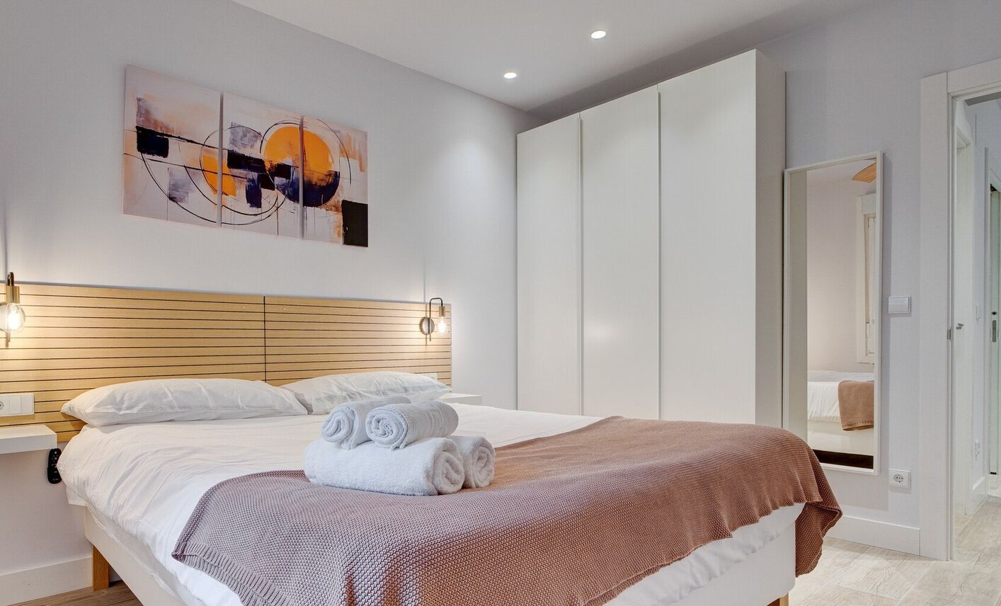A modern bedroom with a wooden headboard, a large bed with white linens, a brown throw blanket, and a wall-mounted abstract painting.