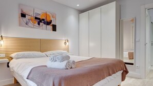 A modern bedroom with a wooden headboard, a large bed with white linens, a brown throw blanket, and a wall-mounted abstract painting.