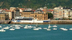 A marina with numerous boats docked, a large white building with a blue roof, and a hillside in the background.