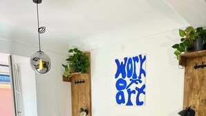 Une chambre avec une tête de lit bleue recouverte de tissu, un lit avec une literie blanche, une œuvre d’art accrochée au mur sur laquelle on peut lire « WORK ART », et une étagère en bois avec des plantes.